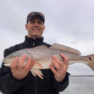 Man holding a large silver fish by the water on an overcast day