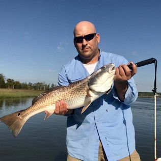 Man holding a large fish on a boat, wearing sunglasses and a blue shirt by the water.