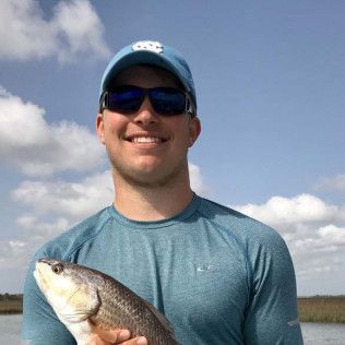 Smiling person in sunglasses and blue cap holding a fish outdoors by the water