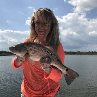 Person in a coral shirt holding a large fish by the water under a cloudy sky