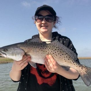 Person holding a large spotted fish outdoors by the water
