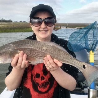 Person holding a large fish outdoors near water, wearing sunglasses and a black cap, smiling.