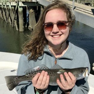 Person smiling while holding a spotted fish on a boat by the water