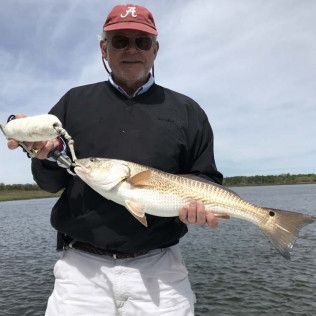 Man holding a large fish on a boat, wearing a red cap and sunglasses on a calm body of water.