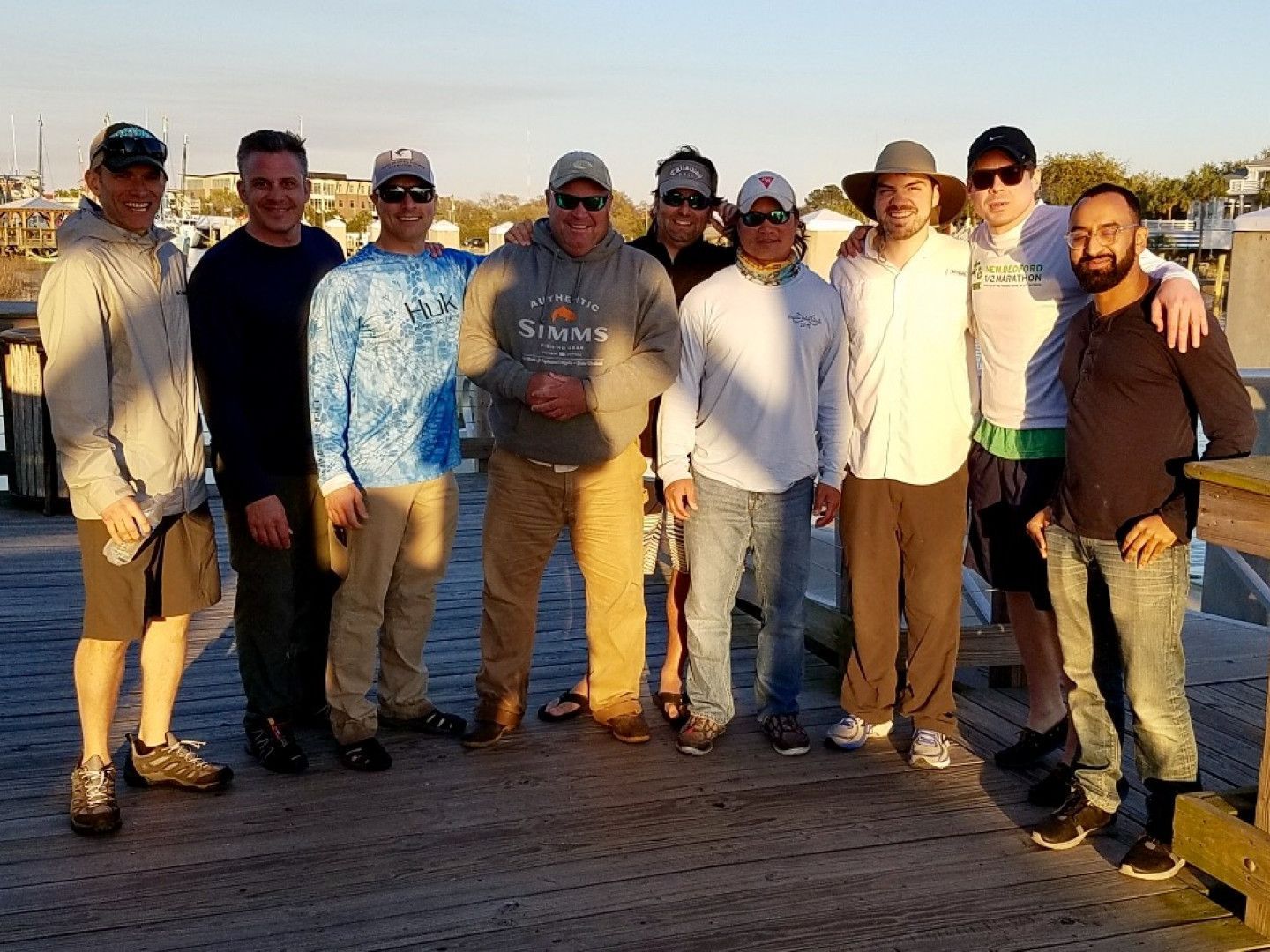 Group of people standing together on a wooden pier at sunset, smiling in casual clothes.