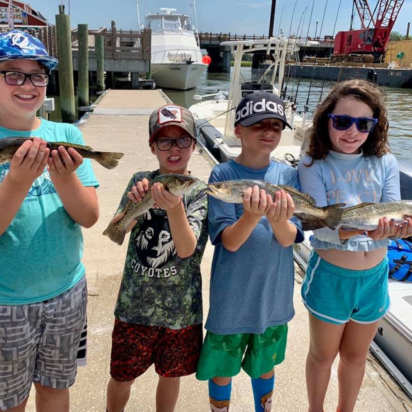 Four children on a dock holding fish, with boats and harbor equipment in the background.