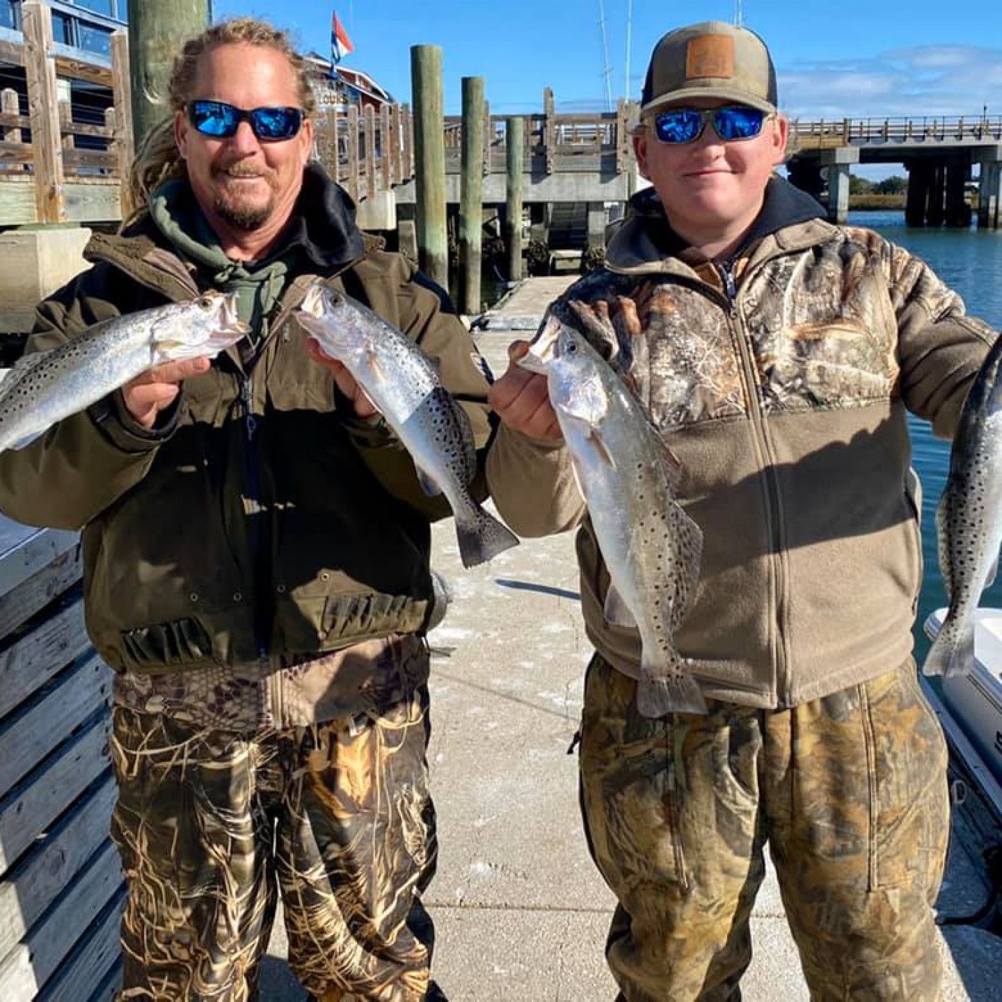 Two anglers in camouflage hold up large fish on a sunny dock by the water