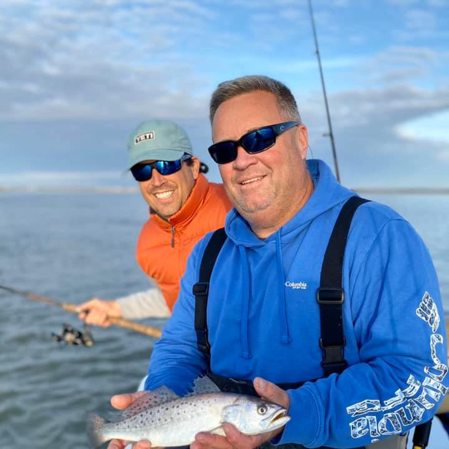 Two smiling anglers on a boat, one holding a fish, with fishing rods and open water behind them
