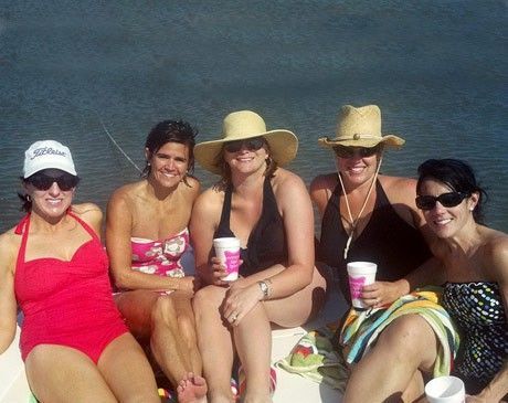 Five friends sitting on a beach, smiling in swimwear and sun hats, holding drinks.