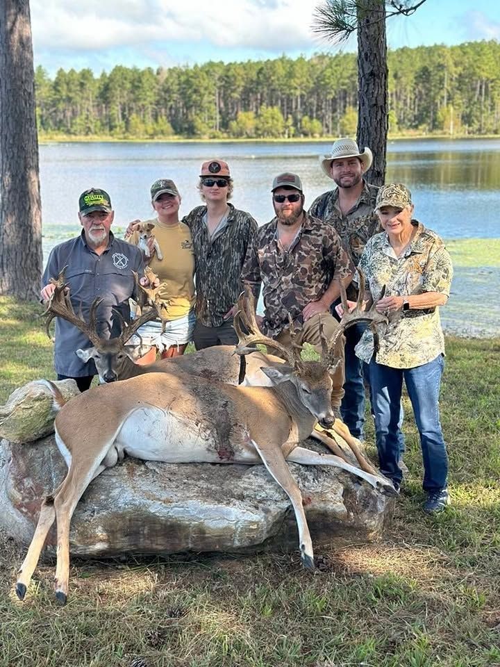 Five hunters posing beside a deer near a lakeshore with forested hills in the background