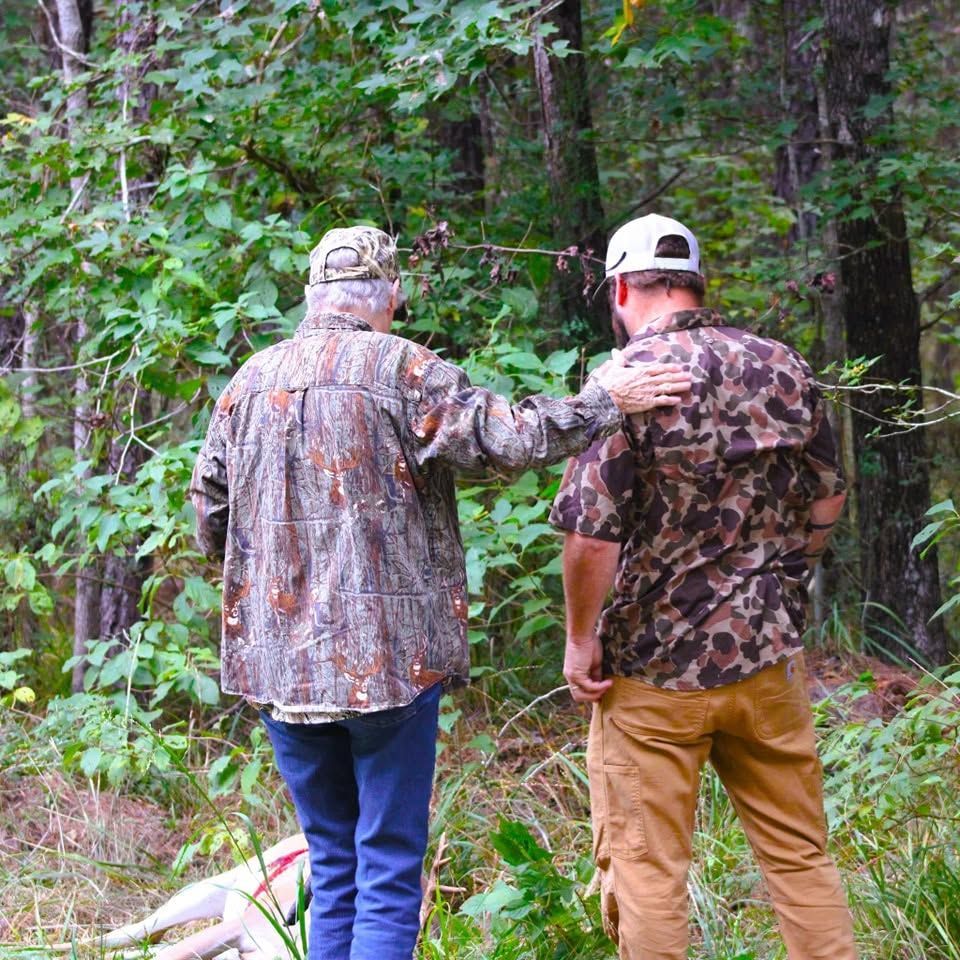 Two hunters in camo walking through a wooded area, one resting a hand on the other's shoulder.