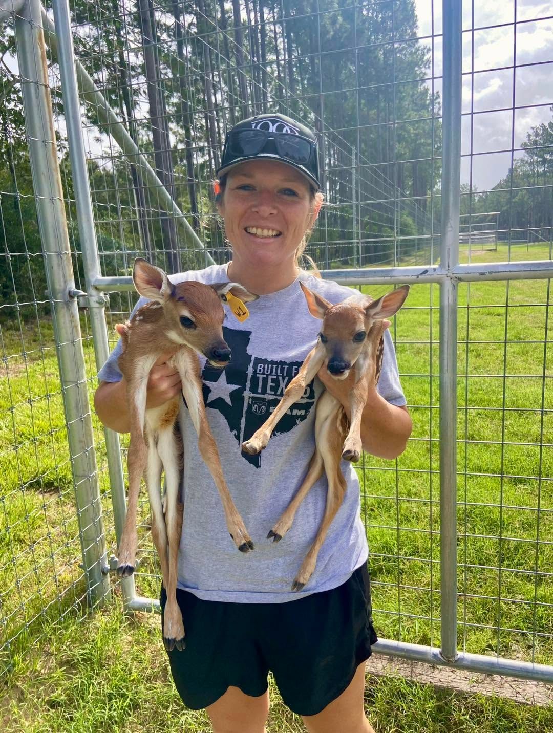 Person smiling and holding two small dogs in front of a fenced grassy field