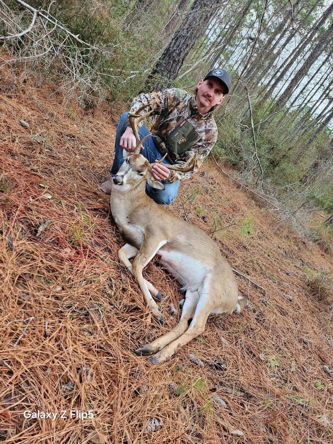 Hunter kneels beside a deer on a forest floor covered in pine needles, holding its head.