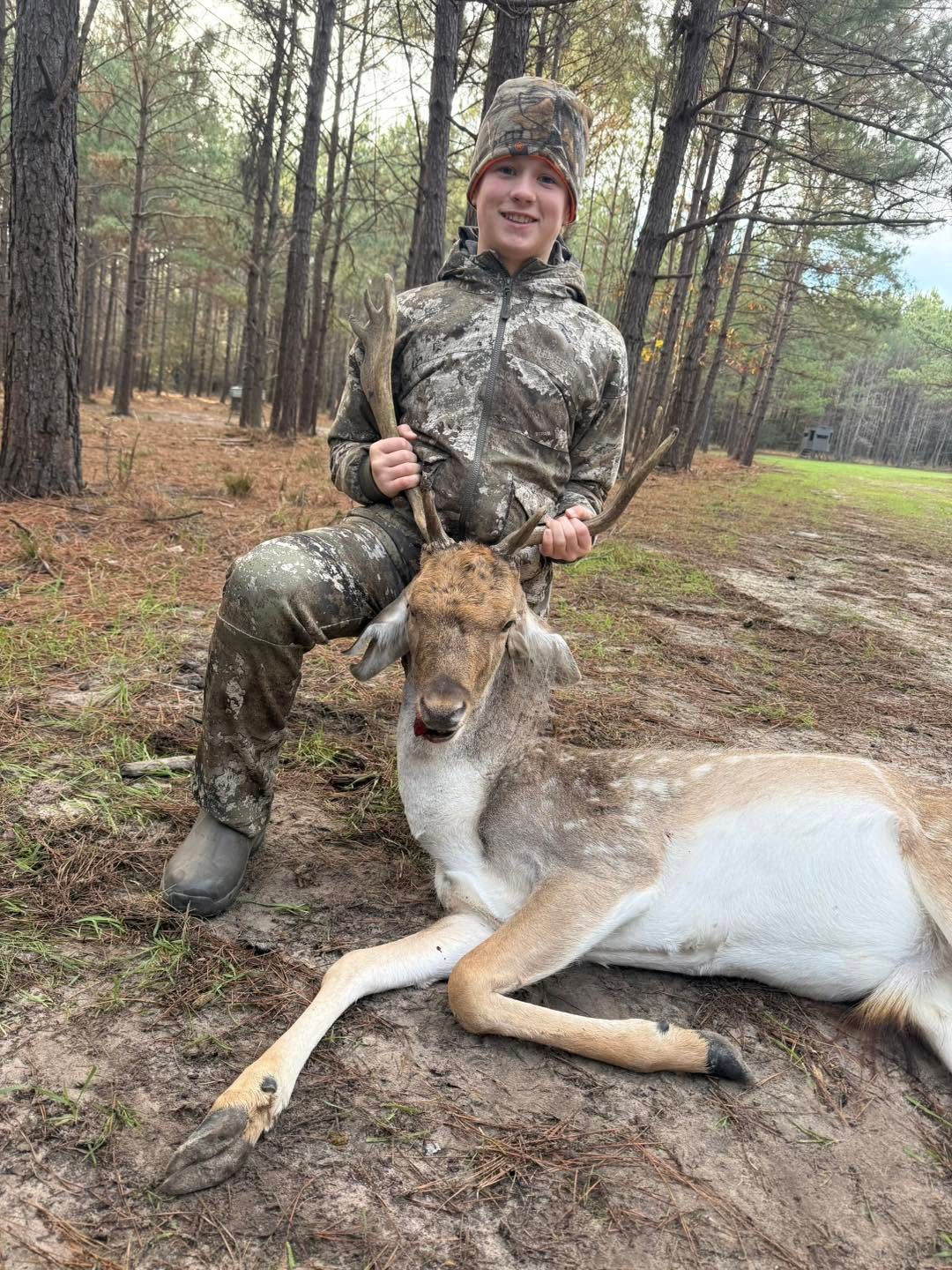 Hunter in camouflage kneels beside a deer in a wooded field.
