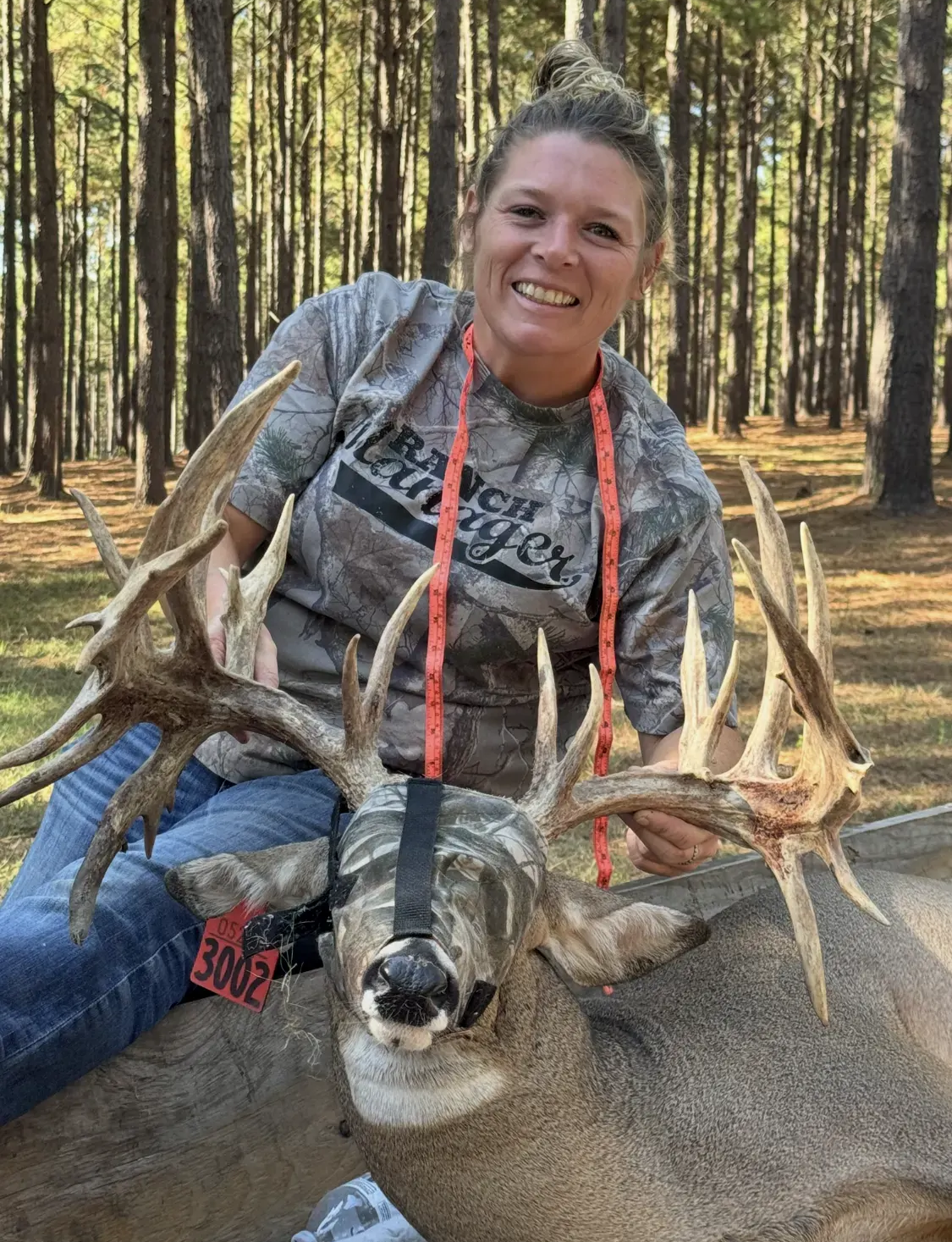 Woman posing with a large buck in a pine forest, smiling with antlers prominent.