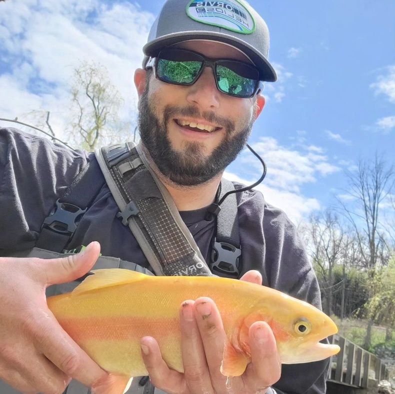 David Kuhn, Founder of The Rods for Wheels Organization, smiling while holding a bright yellow-orange trout outdoors against a blue sky.