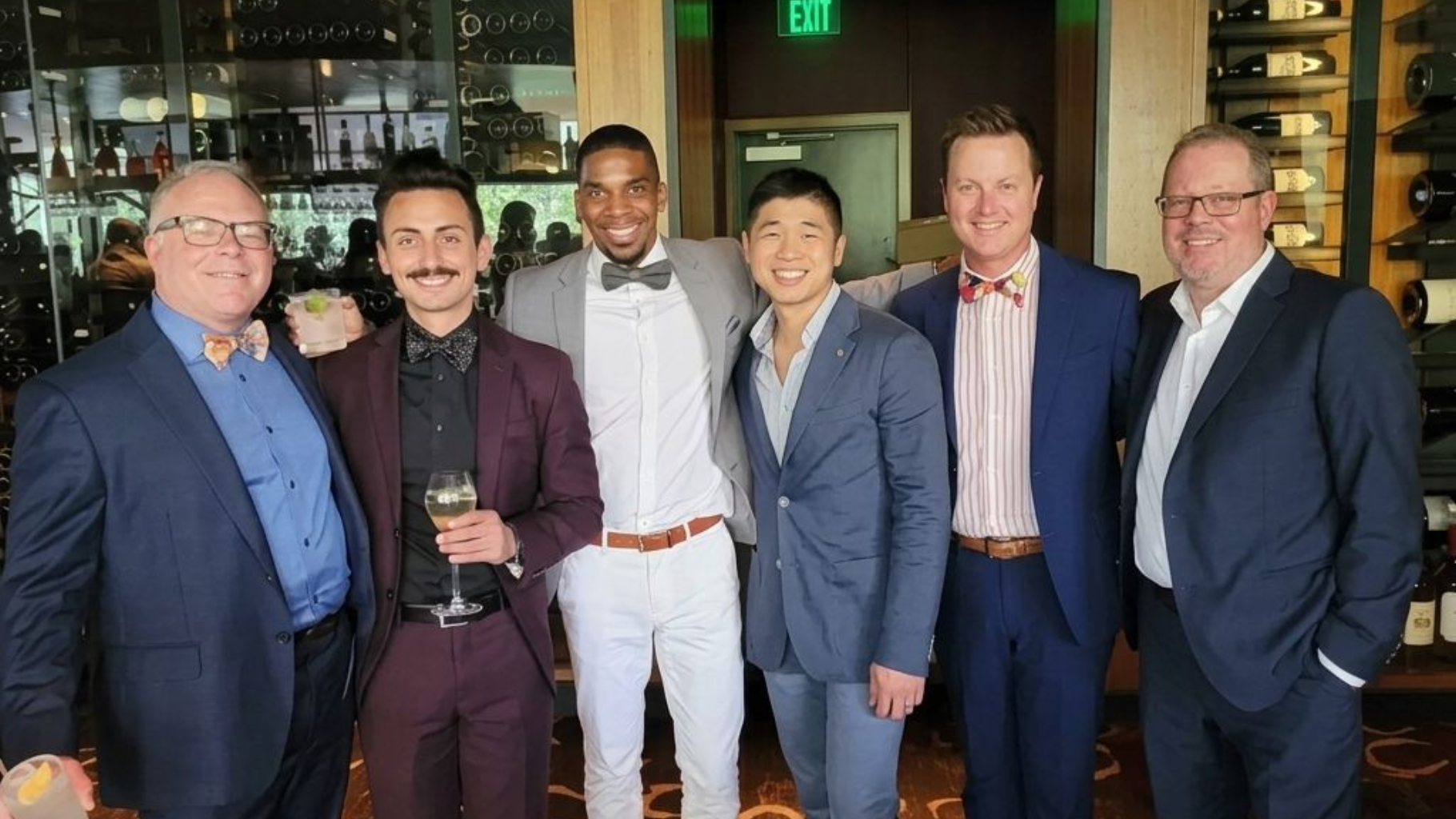 Six people in suits posing together indoors at a social event, smiling and holding drinks