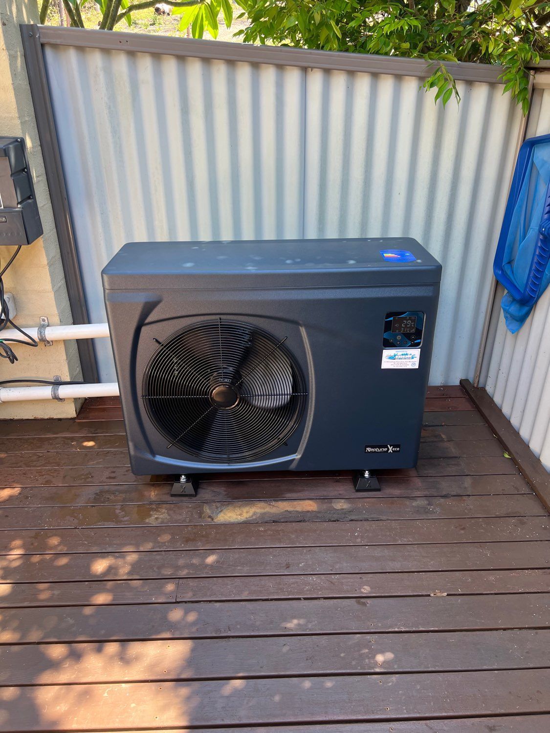 Outdoor air conditioning unit on a wooden deck beside a corrugated fence, with pipes and a blue cloth nearby— Cascade Pool & Spa Centre in Port Macquarie, NSW