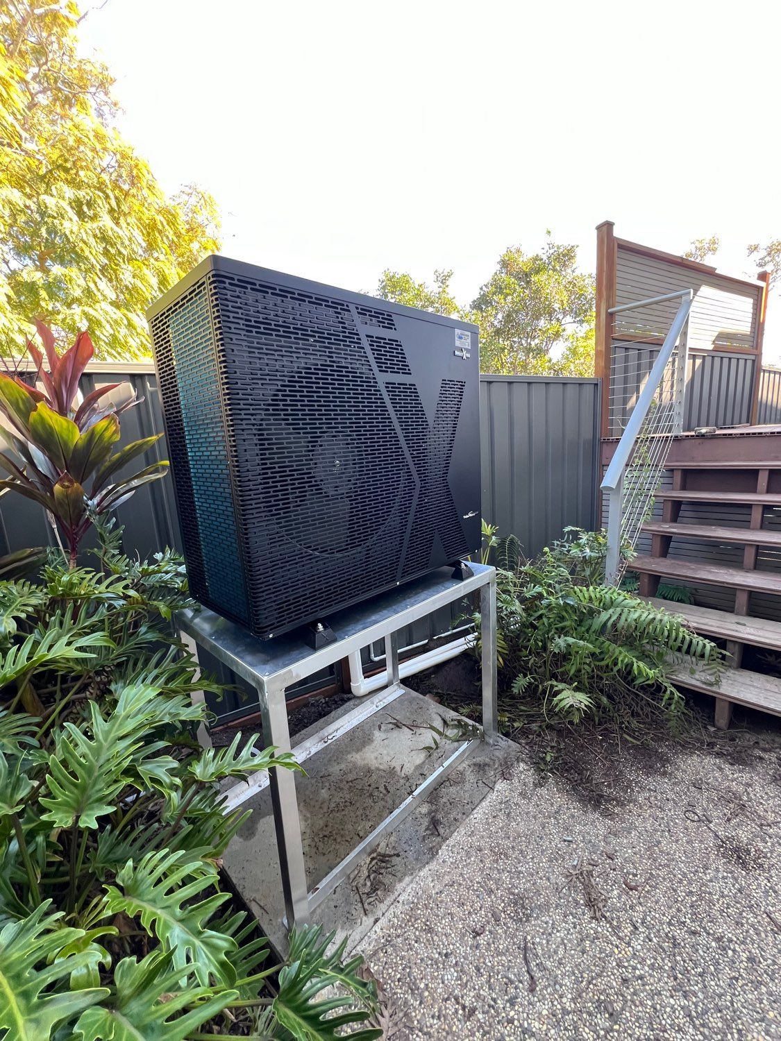 Black outdoor air-conditioning unit beside a fence and stairs in a gravel garden — Cascade Pool & Spa Centre in Port Macquarie, NSW
