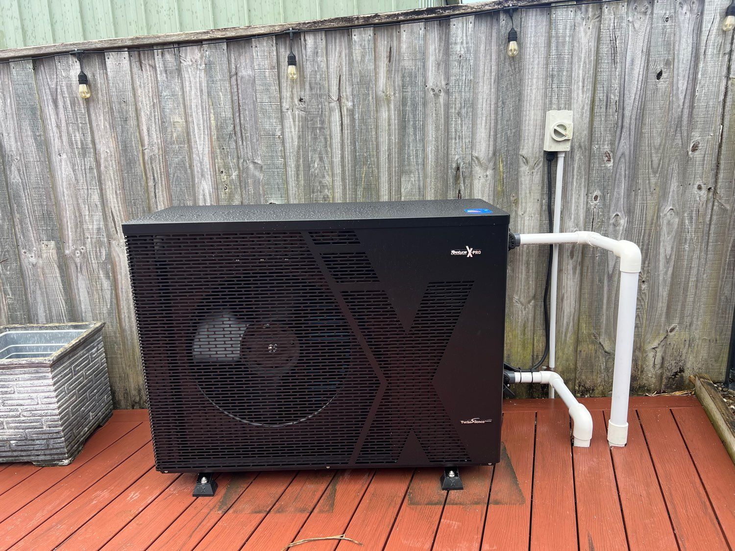 Black outdoor HVAC unit beside white PVC pipes against a weathered wooden fence. — Cascade Pool & Spa Centre in Port Macquarie, NSW