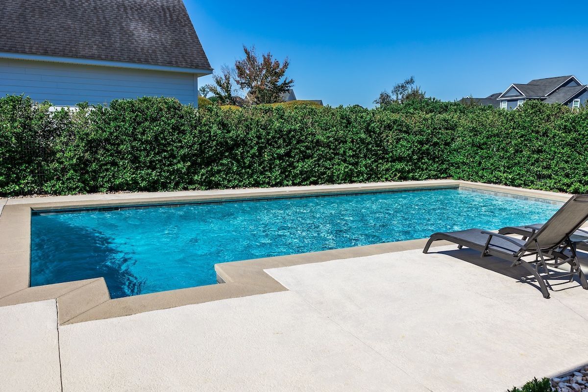 Rectangular pool with turquoise water and light stone patio, bordered by green bushes. A lounge chair sits poolside — Cascade Pool & Spa Centre in Port Macquarie, NSW