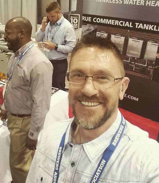 A selfie of a smiling man in a light shirt with a conference lanyard, with two people in the background at a trade show.