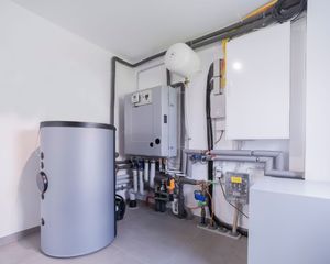 A utility room featuring a cylindrical gray water tank, a central gray heat pump unit, and white wall-mounted equipment.