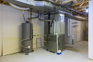 A gray water heater and a furnace located in a basement with exposed joists and a white concrete floor.