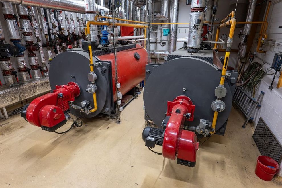 Two large industrial boilers with red burner units installed in a mechanical utility room with pipes and insulated tanks.