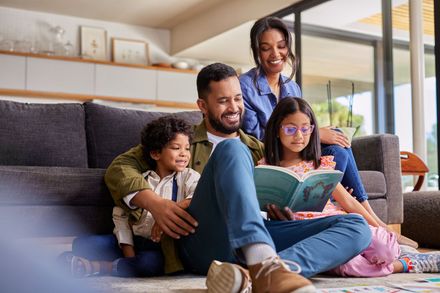 A family sits together on a living room floor, with the parents helping their two children read a book.