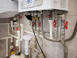 A wall-mounted gas boiler with exposed white piping, metal valves with red handles, and a drainage funnel in a tiled room.