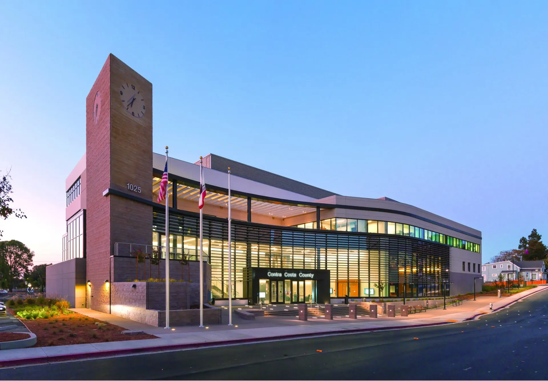 Modern glass office building at dusk with a brick tower and illuminated interior lights