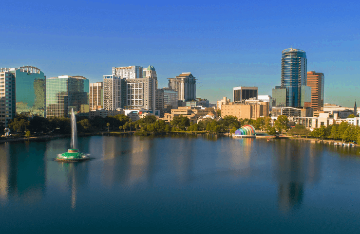 City skyline across a calm lake at sunset, with modern high-rises and a sailboat in the water