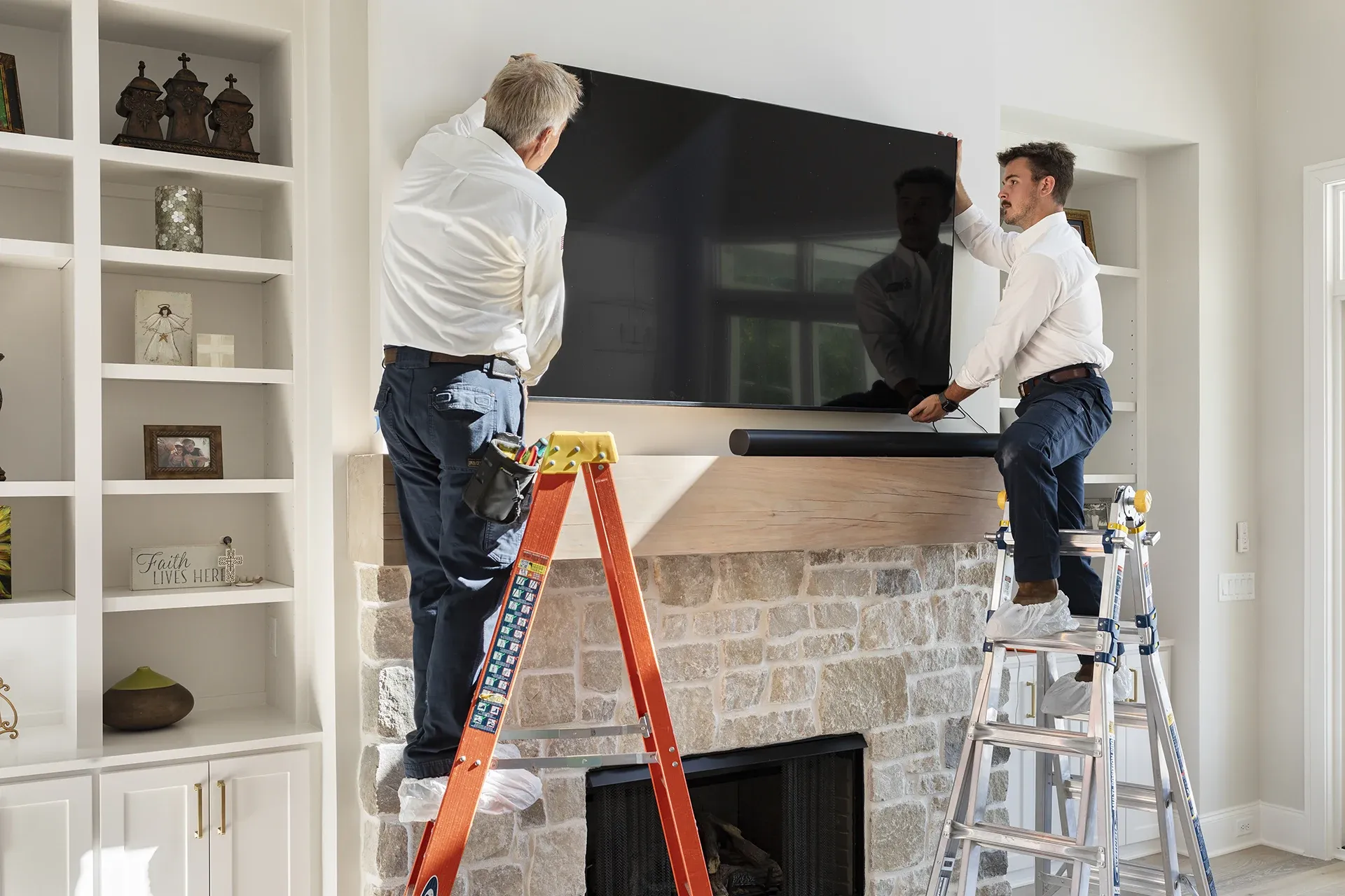 Two people installing a TV above a fireplace, using ladders.