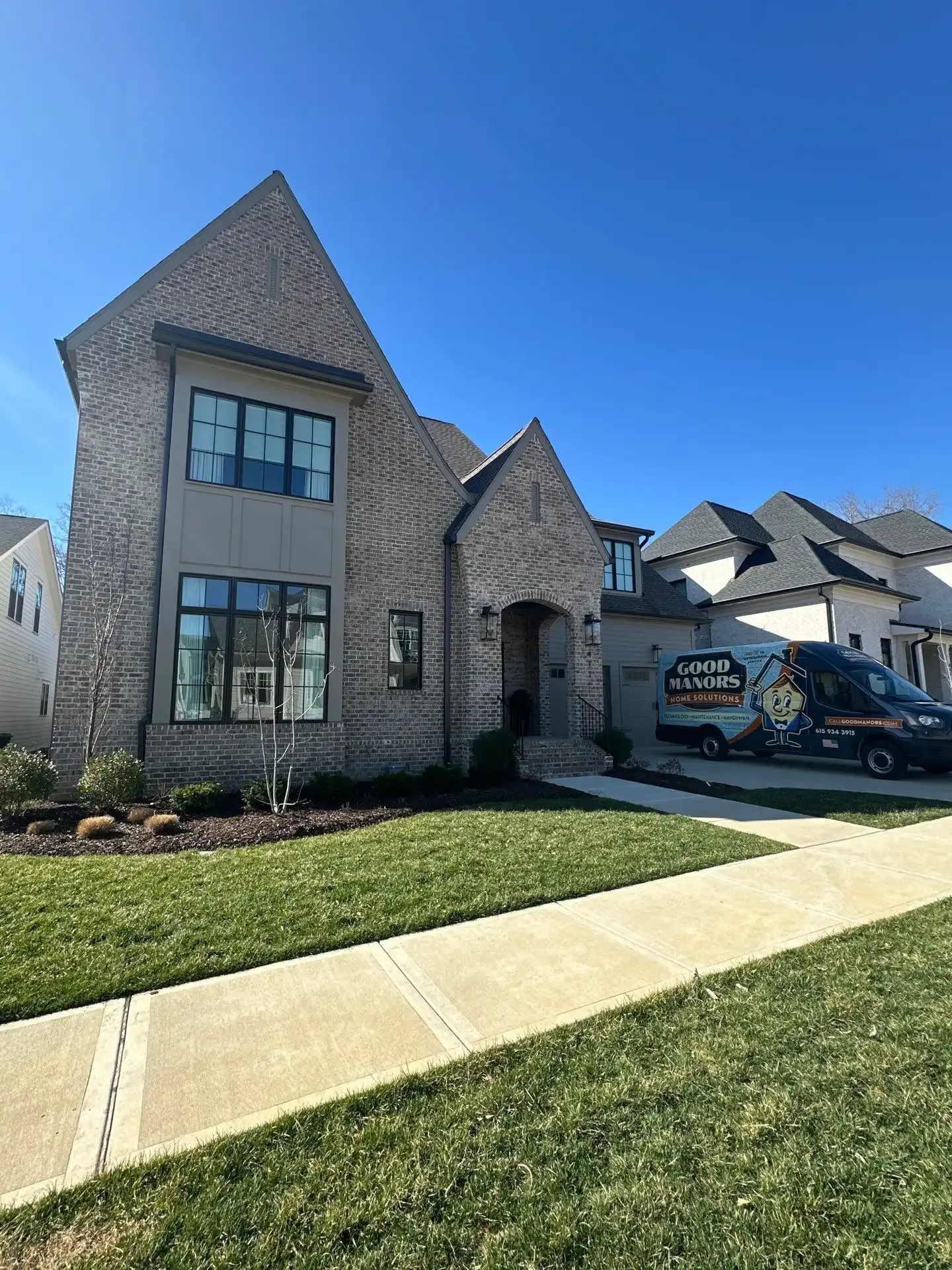 Two-story brick house with dark window frames; parked van on the right. Blue sky and green grass.