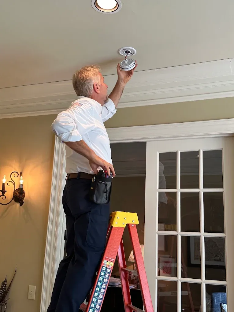 Man on a ladder installs a smoke detector on a ceiling.