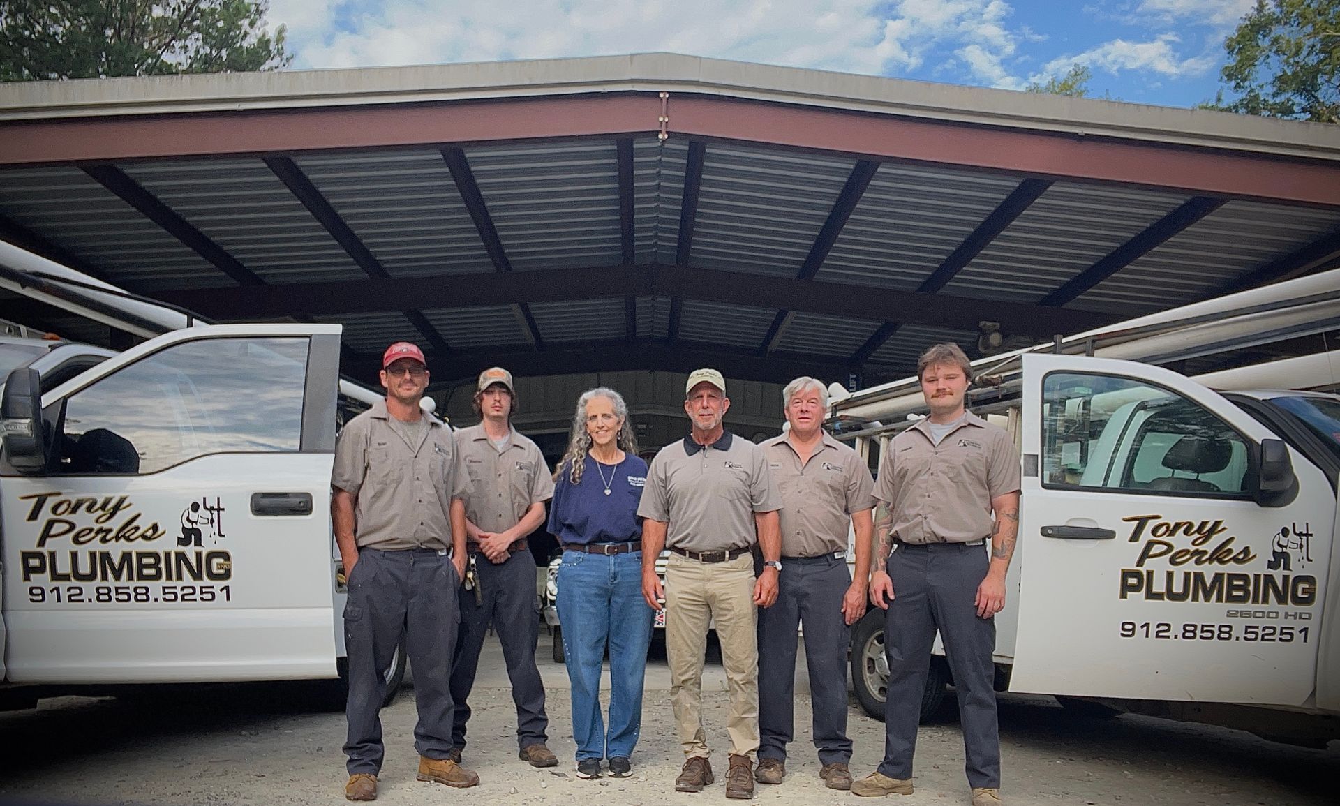 Five plumbing company employees standing in front of white service vans under a metal awning