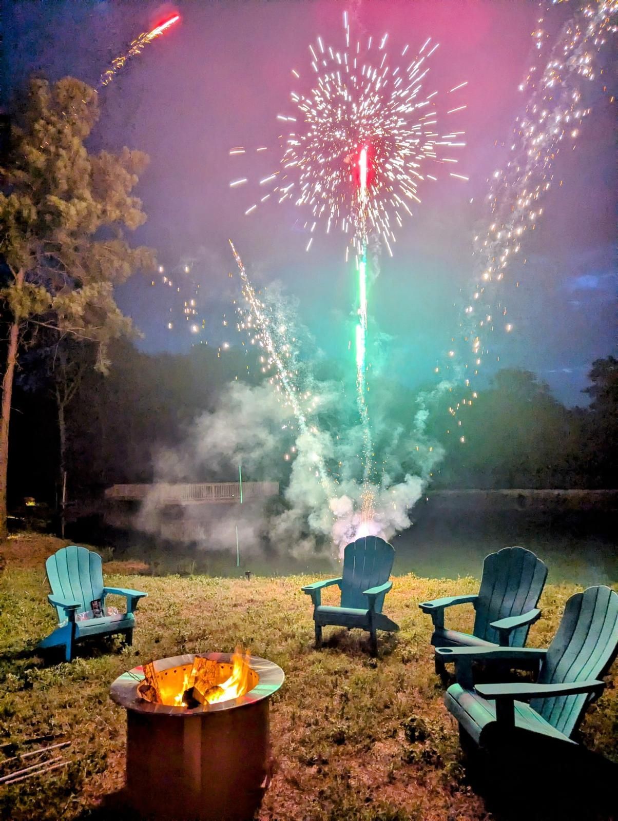 Firework display over a backyard with Adirondack chairs around a fire pit, trees, and a pond at dusk.