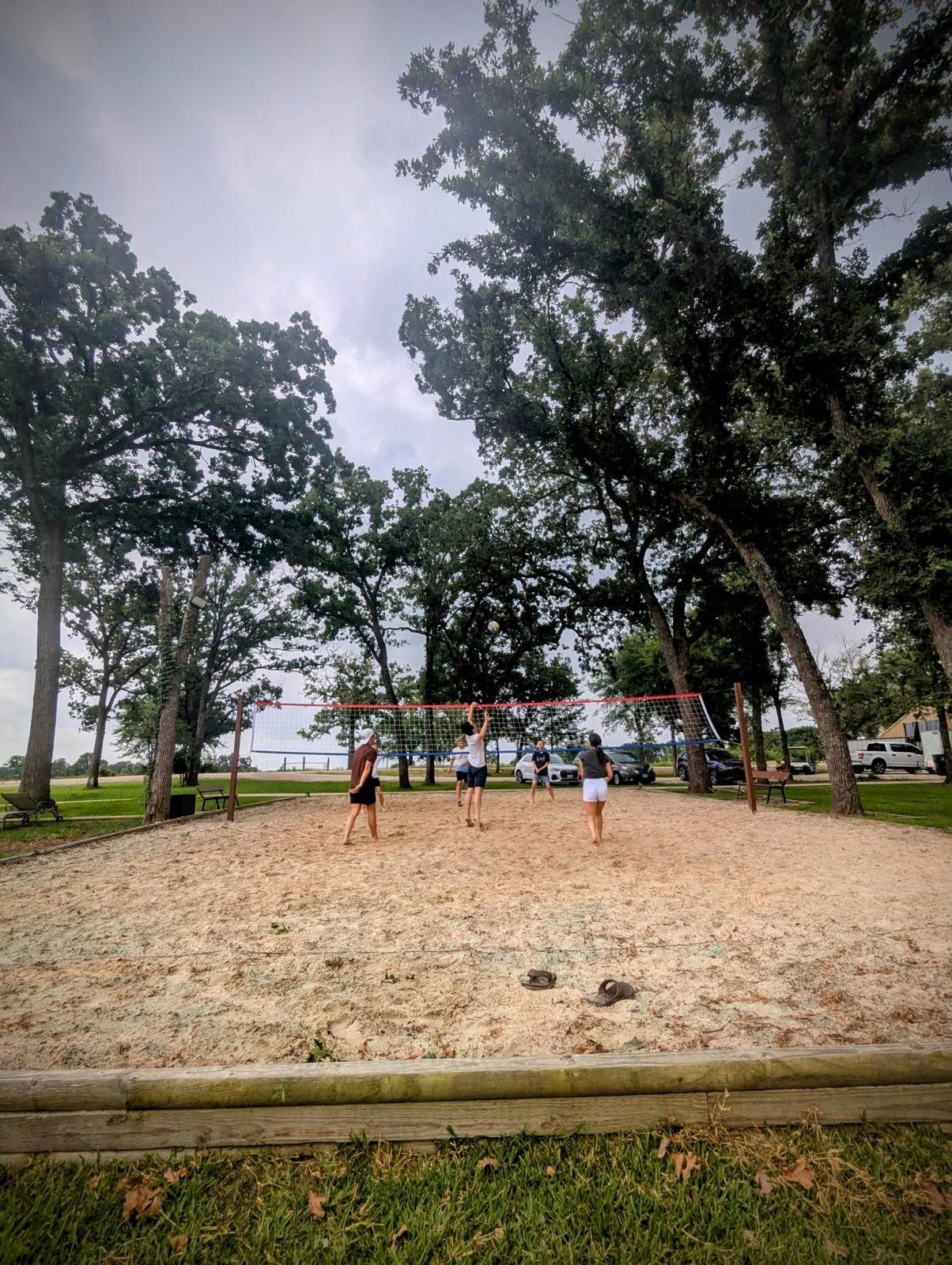 Beach volleyball game in progress, sandy court surrounded by grass and trees under a cloudy sky. People are playing.