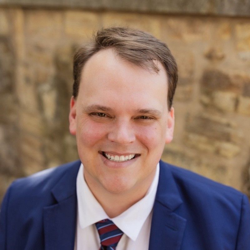Smiling man in a blue suit and striped tie in front of a stone wall