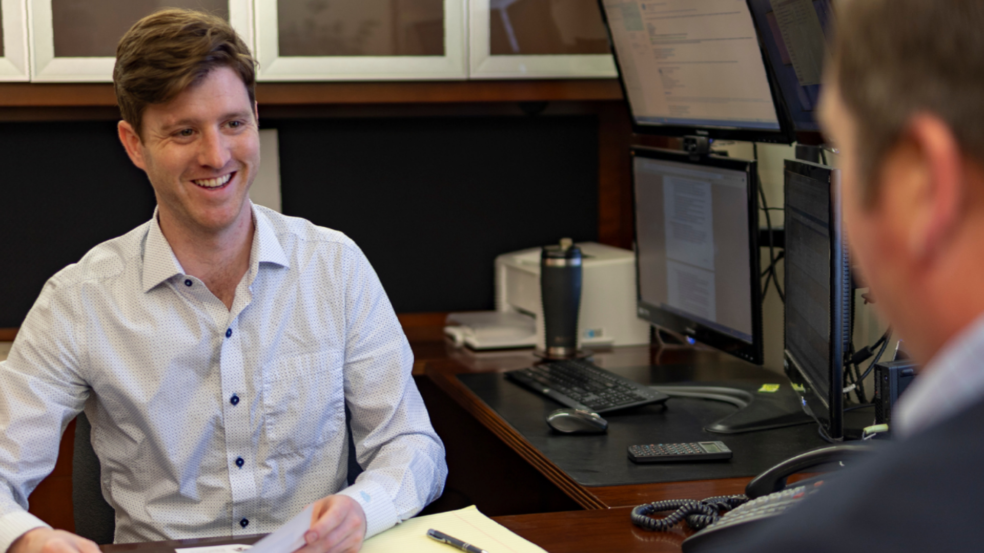 Man smiling during a meeting across a desk in an office, with computer monitors in the background