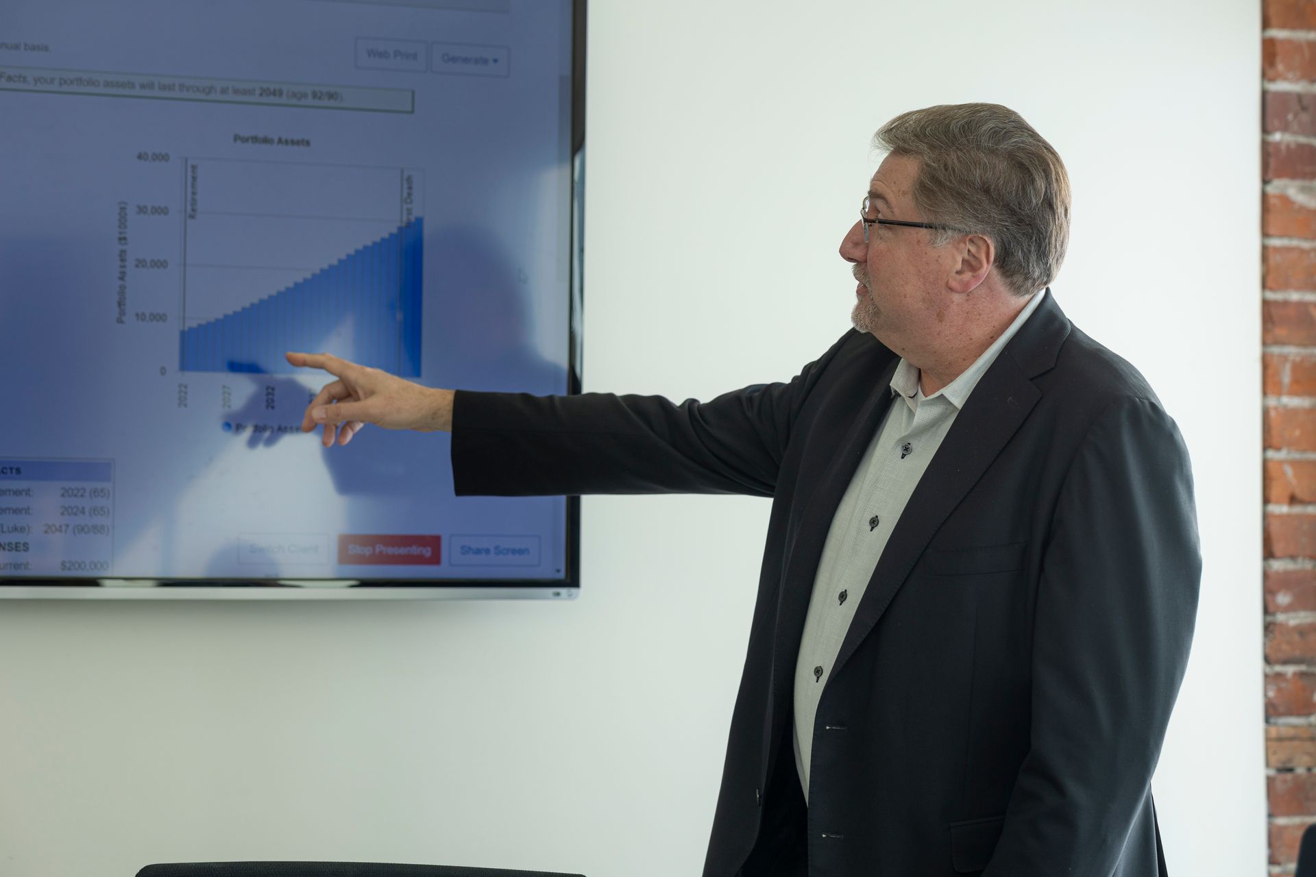 Man in a dark suit points to a blue chart on a wall-mounted screen during a presentation