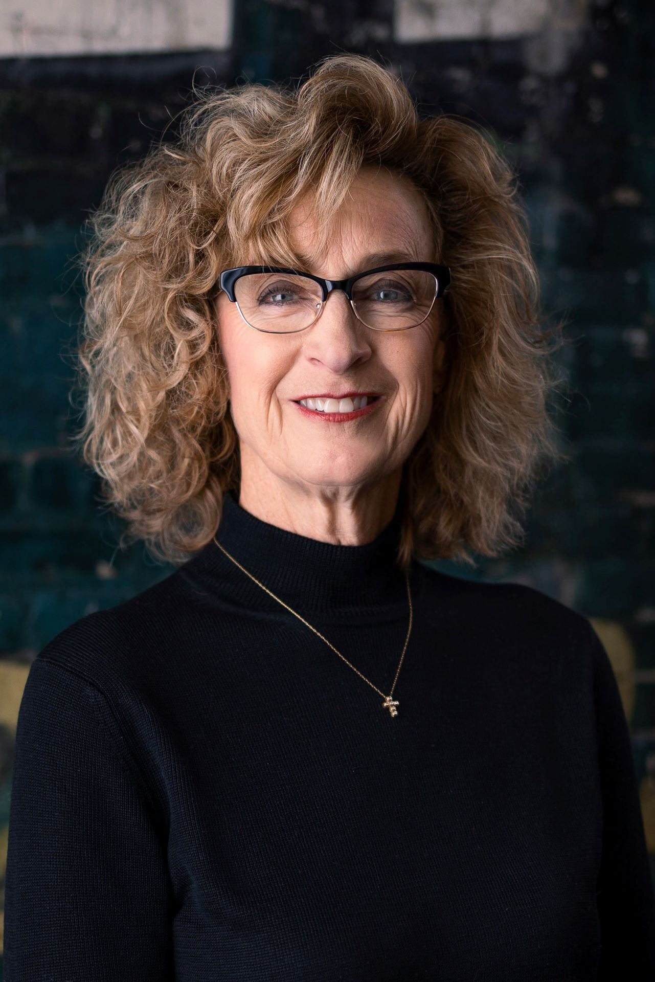 Portrait of a smiling woman with curly hair and glasses, wearing a black top against a dark background