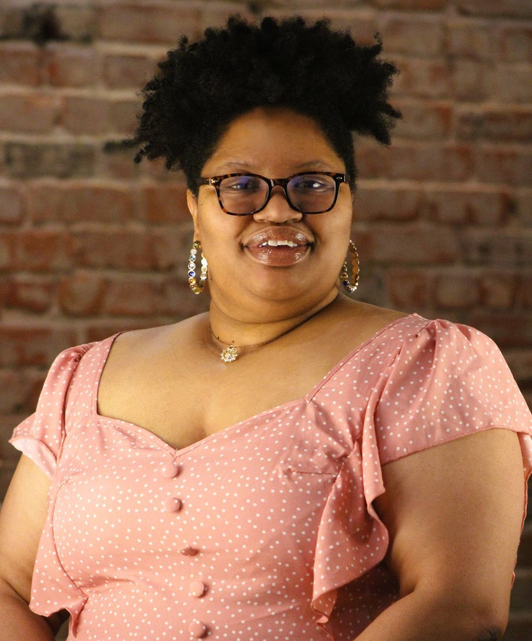 Smiling person in a pink polka-dot dress against a brick wall