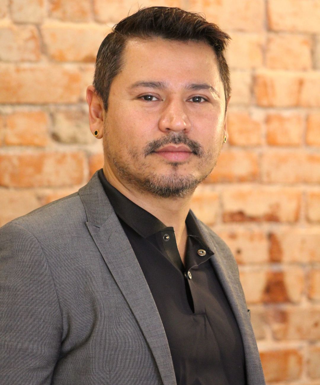 Man in a gray blazer and black shirt posed against a brick wall background