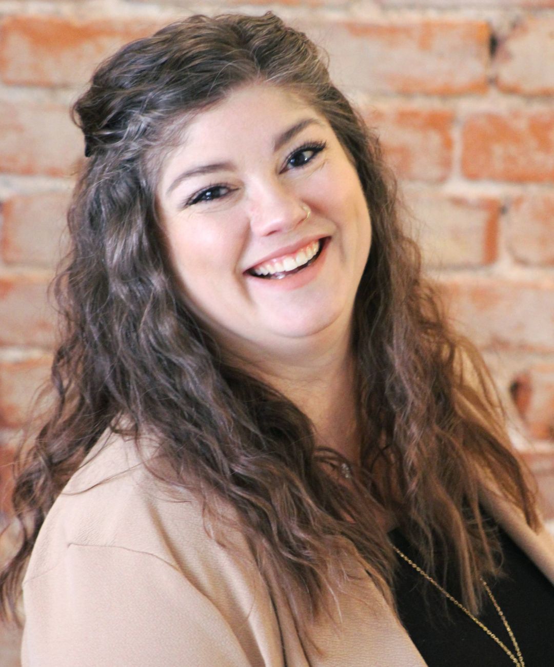 Smiling person with long wavy hair in a tan jacket against a brick wall.
