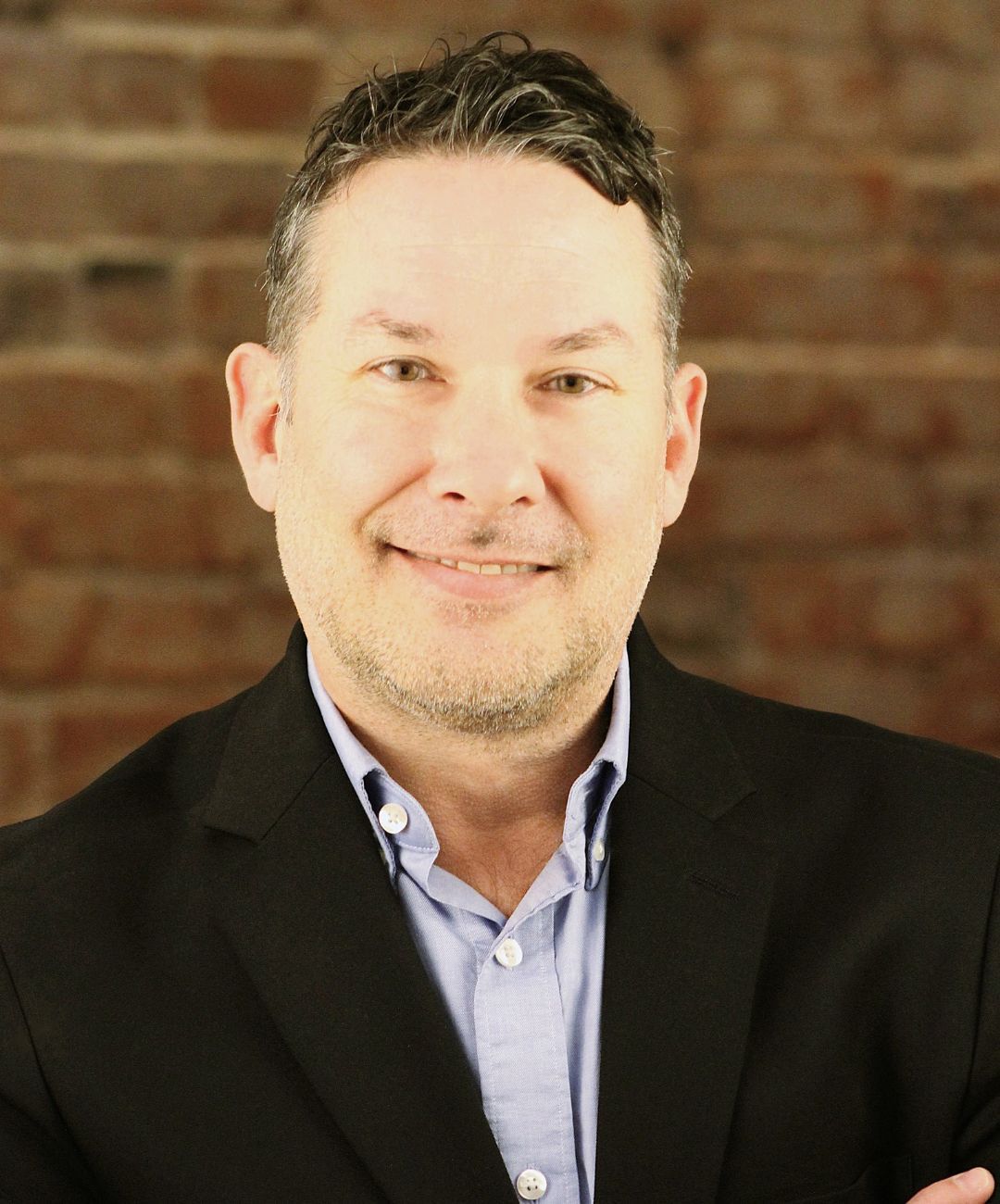 Portrait of a smiling person in a black blazer and light blue shirt against a brick wall background