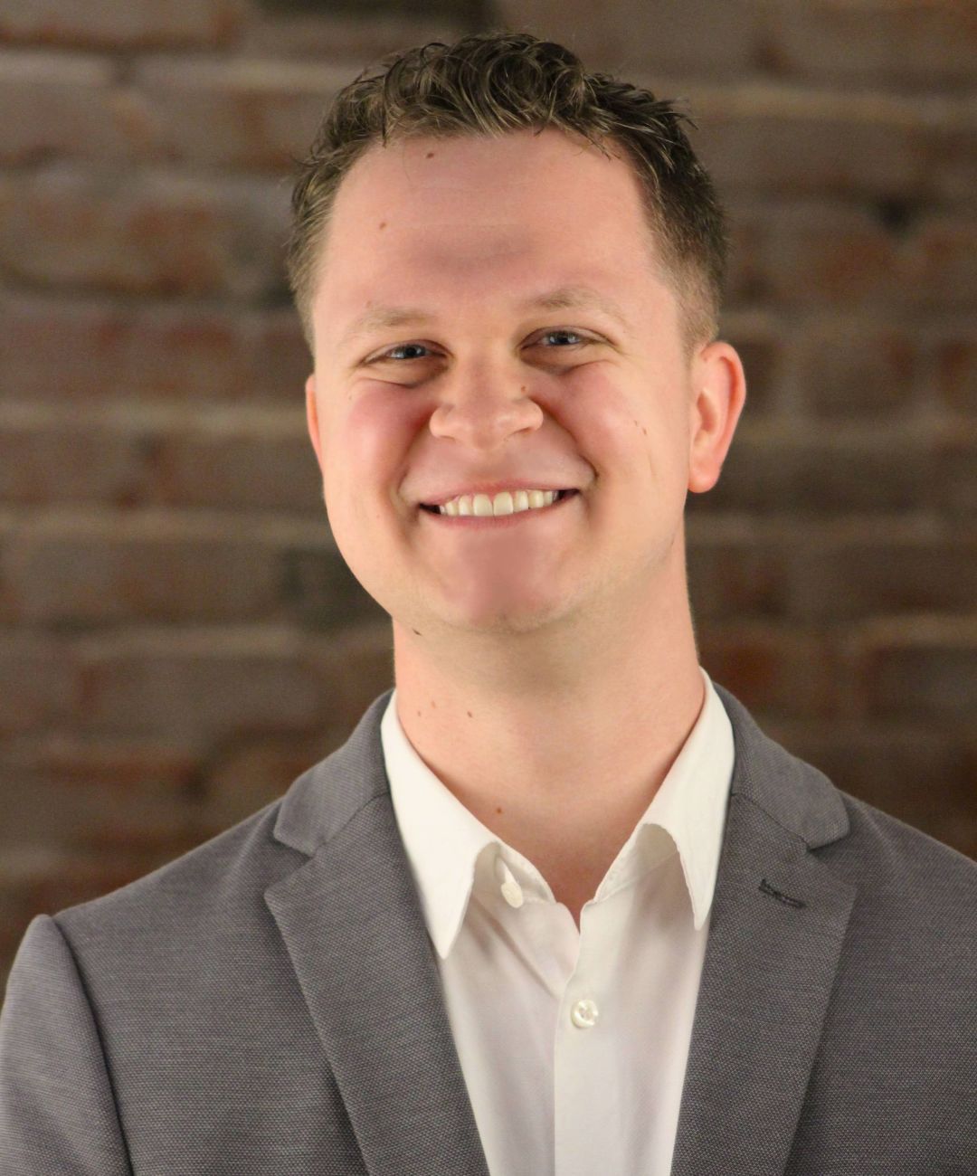 Smiling man in a gray blazer and white shirt against a brick wall background