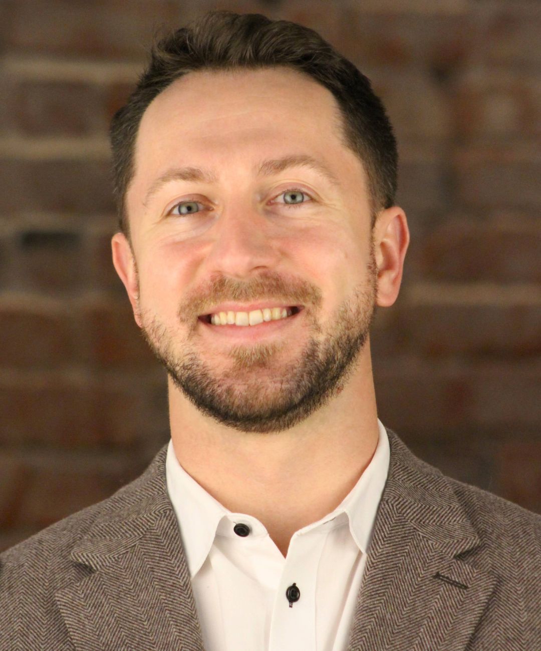 Smiling man in a gray blazer and white shirt against a brick wall background