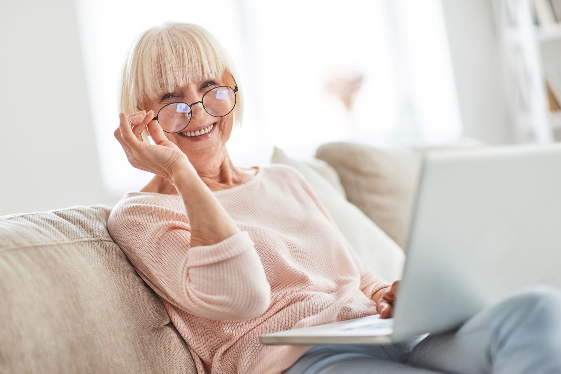 Smiling woman with glasses using a laptop on a couch in a bright living room